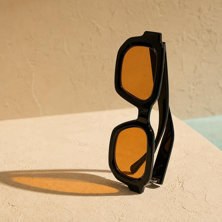 Gafas de sol negras con lente ámbar proyectando luz naranja sobre una pared de arena